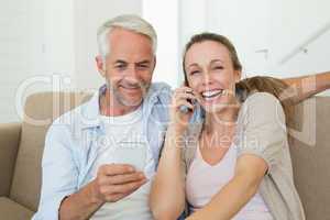 Happy couple sitting on couch talking and texting on their phone