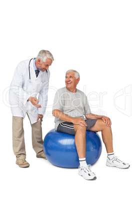 Male therapist looking at senior man sit on exercise ball