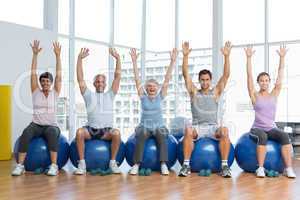 Class sitting on exercise balls and raising hands in gym