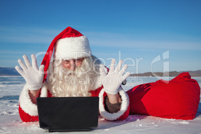 Santa Claus enjoys lying in the snow, looking at laptop news