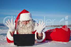 Santa Claus enjoys lying in the snow, looking at laptop news