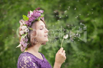 Cute girl in a wreath of lilacs blowing on a dandelion