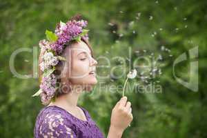 Cute girl in a wreath of lilacs blowing on a dandelion