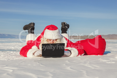 Santa Claus lying on the snow, looking at laptop news