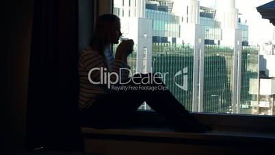Woman drinking coffee on the windowsill with city view in window