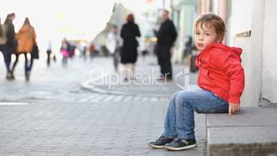 Little boy sitting alone in the street, people passing by