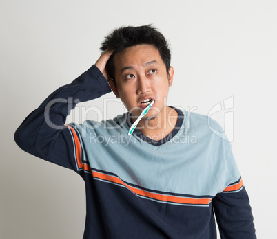 Asian male scratching head and looking up while brushing teeth