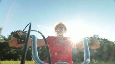 Cheerful senior woman riding bicycle against sun and blue sky POV