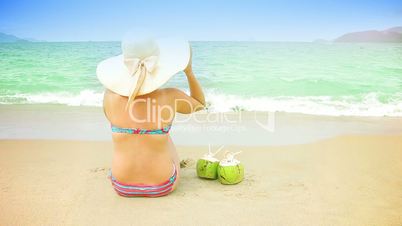 Woman in hat and 2 coconuts sit on a sandy beach.