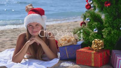 Smiling girl in Santa hat with gift boxes under Christmas tree on the beach