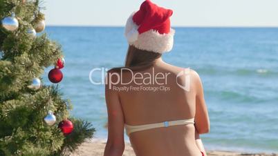 Girl in Santa hat near Christmas tree on the beach rear view