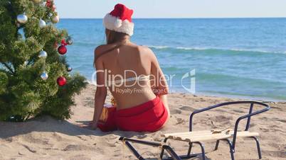 Girl in Santa hat near Christmas tree on the beach rear view