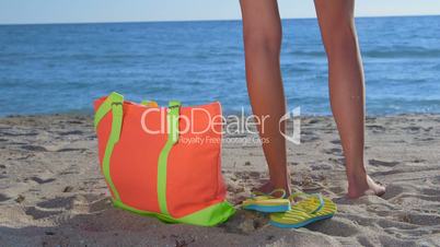 Colorful bag and flip-flops on sandy beach bikini woman in background