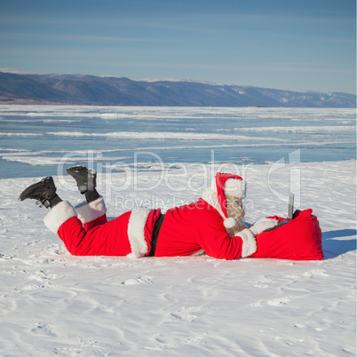 Santa Claus lying on the snow, looking at laptop news