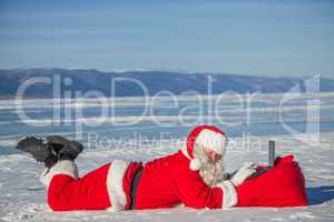 Santa Claus lying on the snow, looking at laptop news