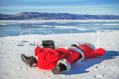 Santa Claus lying on the snow, looking at laptop news