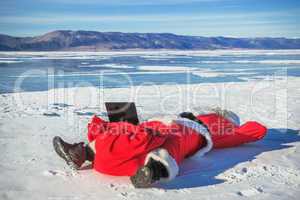 Santa Claus lying on the snow, looking at laptop news