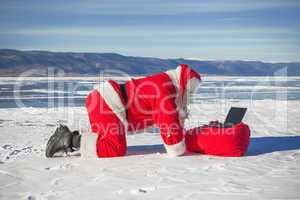 Santa Claus lying on the snow, looking at laptop news