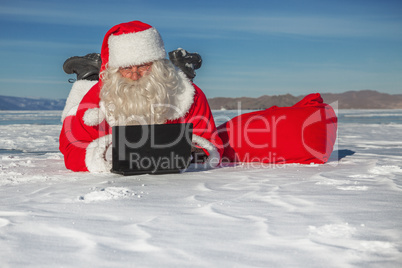 Santa Claus lying on the snow, looking at laptop news