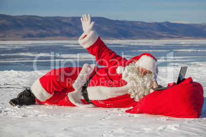 Santa Claus lying on the snow, looking at laptop news