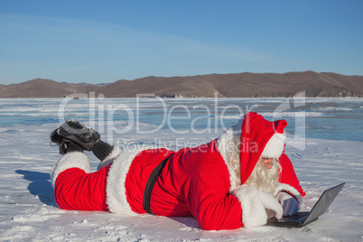 Santa Claus lying on the snow, looking at laptop news