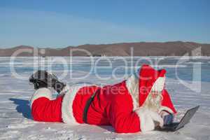 Santa Claus lying on the snow, looking at laptop news