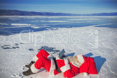 Santa Claus lying on the snow, looking at laptop news