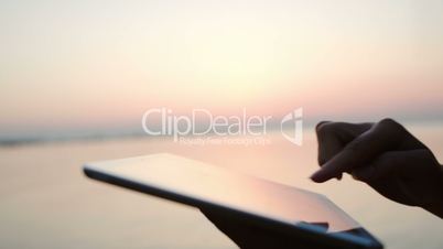 Female hands typing on tablet PC by sea at sunset
