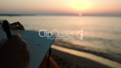 Woman making notes sitting by sea at sunset