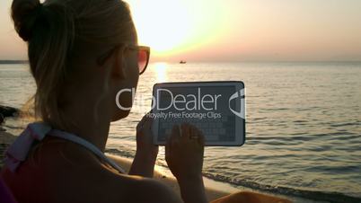 Woman typing on pad sitting by sea at sunset