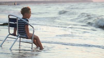 Boy sitting on the chair by sea, waves washing his feet