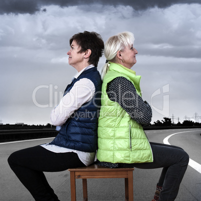 Two women sitting back to back with the chair on the street
