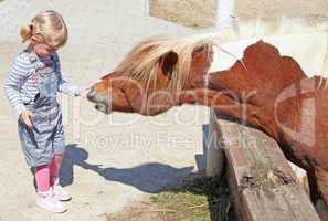 Little blonde girl feeding a horse at the zoo on sunny summer day