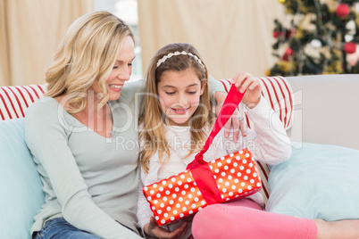Cute little girl sitting on couch opening gift with mum
