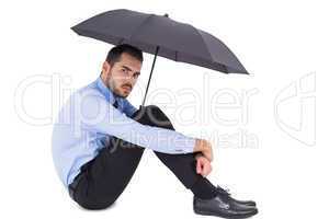 Unsmiling businessman holding umbrella sitting on the floor
