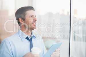 Businessman holding disposable cup and tablet looking out the wi