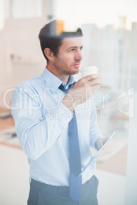 Businessman holding disposable cup and tablet looking out the wi