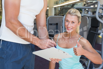 Woman gesturing thumbs up besides trainer with clipboard at gym