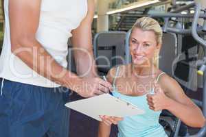 Woman gesturing thumbs up besides trainer with clipboard at gym