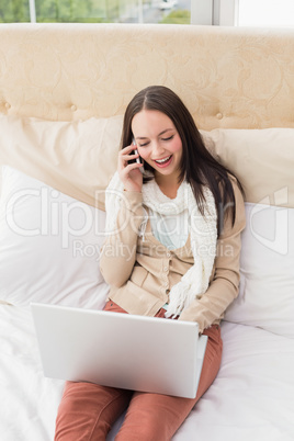 Pretty brunette using laptop on bed making phone call