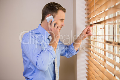 Businessman peeking through blinds while on call