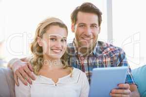 Couple using tablet pc on the couch while looking at camera