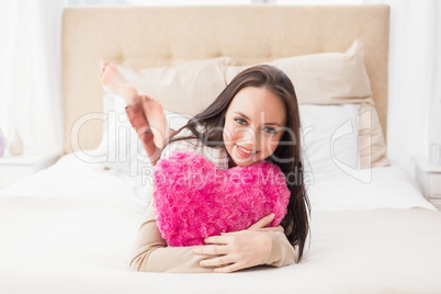 Pretty brunette holding heart cushion on bed