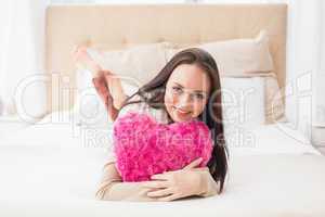 Pretty brunette holding heart cushion on bed