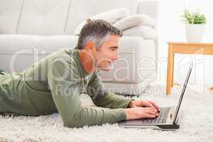 Smiling man lying on carpet using laptop