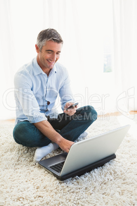 Happy man sitting on carpet using laptop and phone
