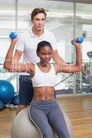 Personal trainer helping client lift dumbbells on exercise ball