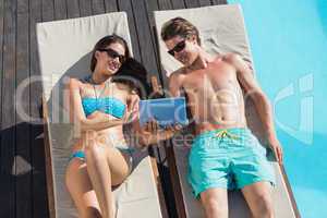 Couple using digital tablet on sun loungers by swimming pool