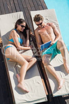 Couple using digital tablet on sun loungers by swimming pool