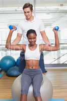 Personal trainer helping client lift dumbbell on exercise ball